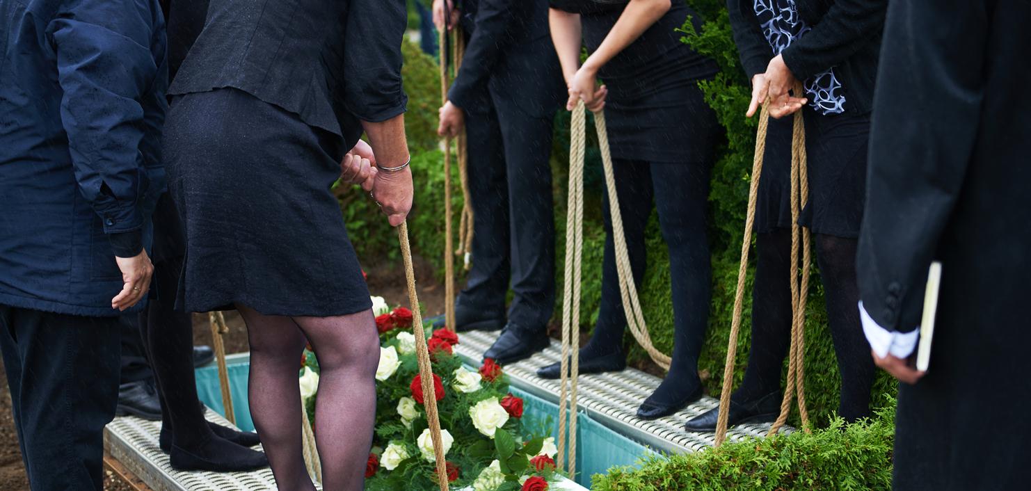 Teilnehmer einer Trauerfeier halten Seile, um Blumenschmuck auf einen Sarg in einer Grabstelle abzusenken.