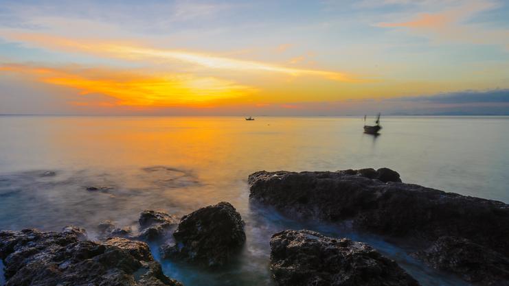 Sonnenuntergang über ruhigem Wasser, Felsen im Vordergrund und ein Boot entfernt auf dem Meer.