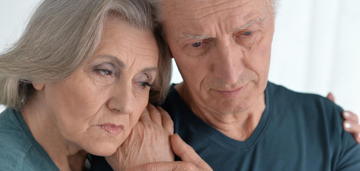 Ein älteres Paar steht eng umschlungen, beide blicken nachdenklich und wirken emotional berührt.