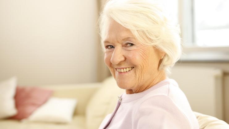 Lächelnde ältere Frau mit grauen Haaren sitzt auf einem Sofa in einem hellen Raum.